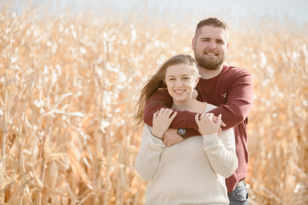 Man standing behind his fiancé both smiling and looking at the camera