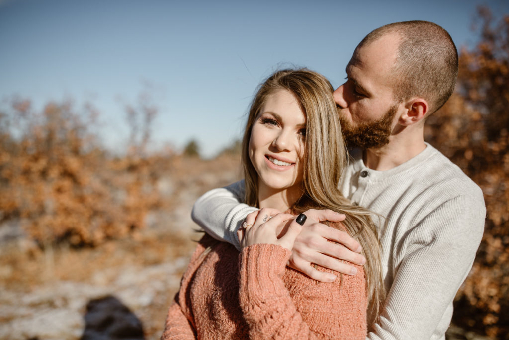 Man standing behind his fiancé, giving her a kiss on the head while she looks at the camera