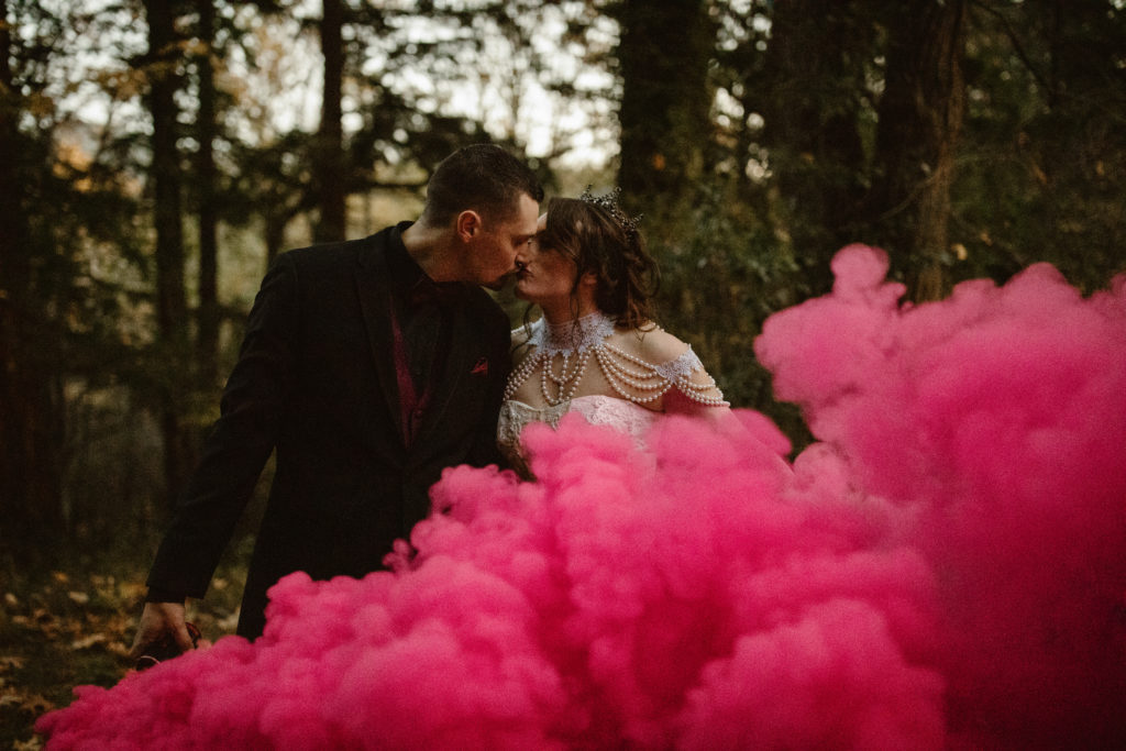 Bride and groom holding hands with colored smoke in front of them.