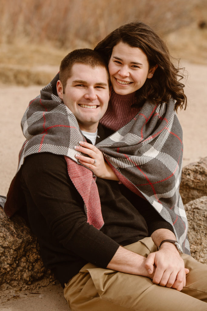 Woman behind her fiancé with her arms around him wrapped in a blanket, both of them smiling