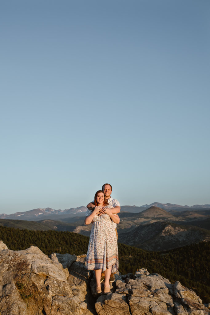 Couple standing on a rock with man standing behind his fiancé with his arms around her