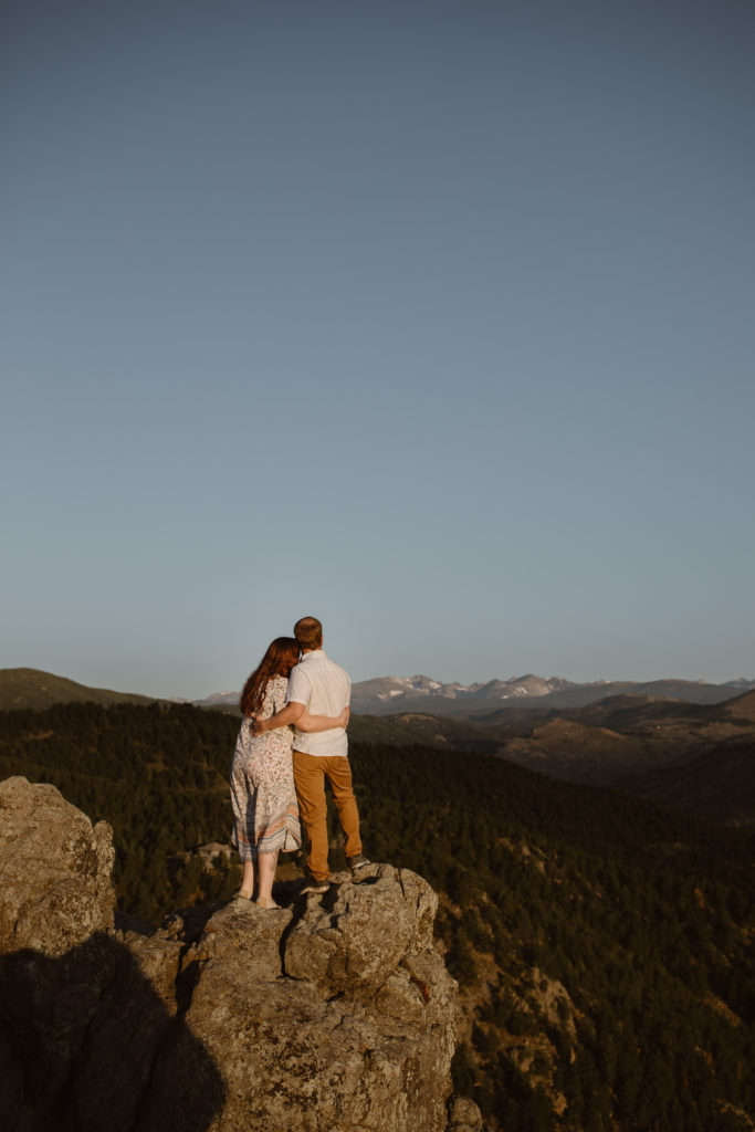 Couple with arms around each other on a rock in the mountains looking at the view