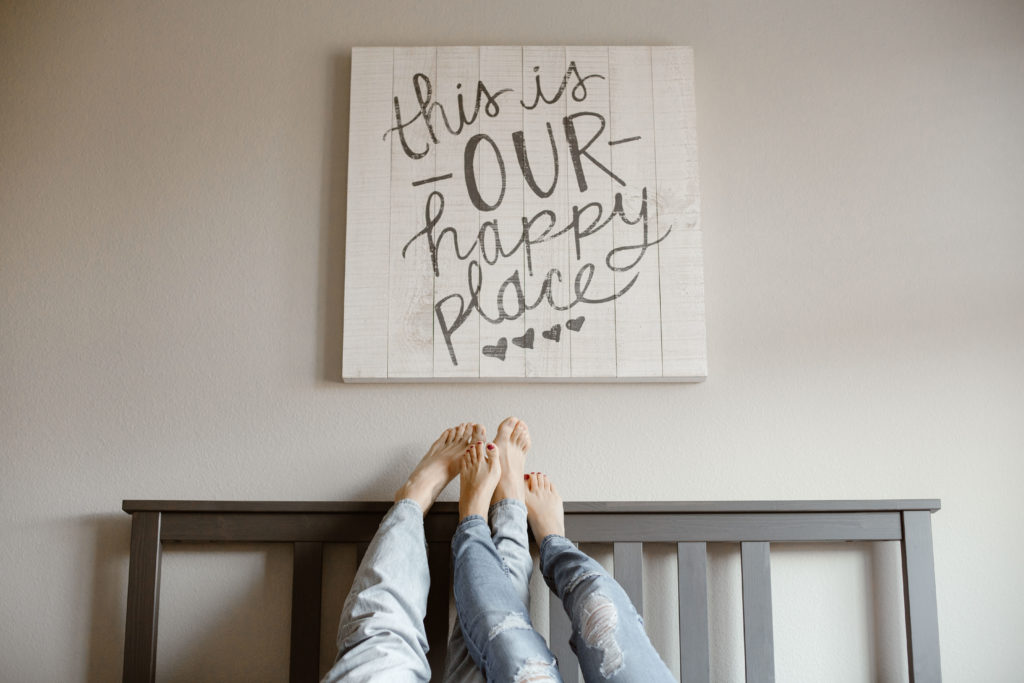 A piece of art saying "this is our happy place" with a couple's feet resting on the wall beneath the art