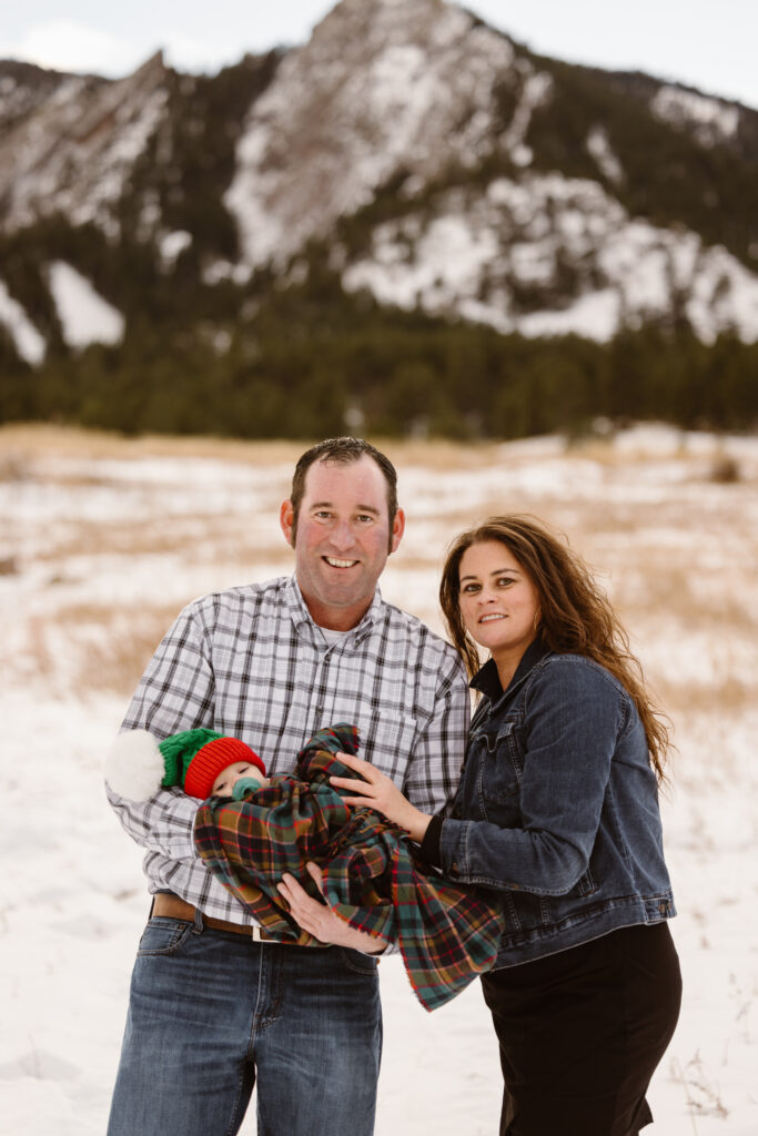 Husband and wife holding their newborn together with snowy Flatirons in the background