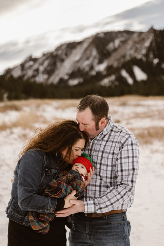 Husband kissing the top of his wife's head and wife kissing their newborn daughter's head with snowy Flatirons in the background