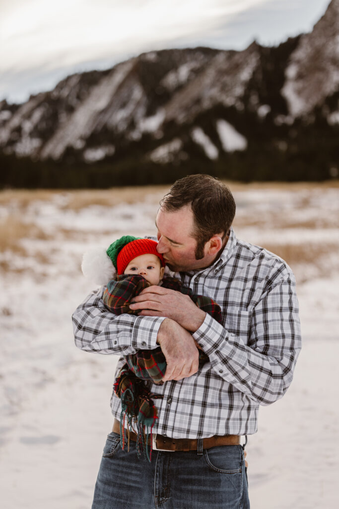 Father kissing the temple of his newborn daughter with snowy Flatirons in the background