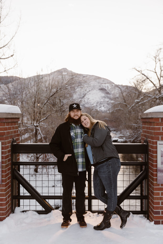 Couple cuddled together on a bridge over a river with a snowy river and mountain in the background