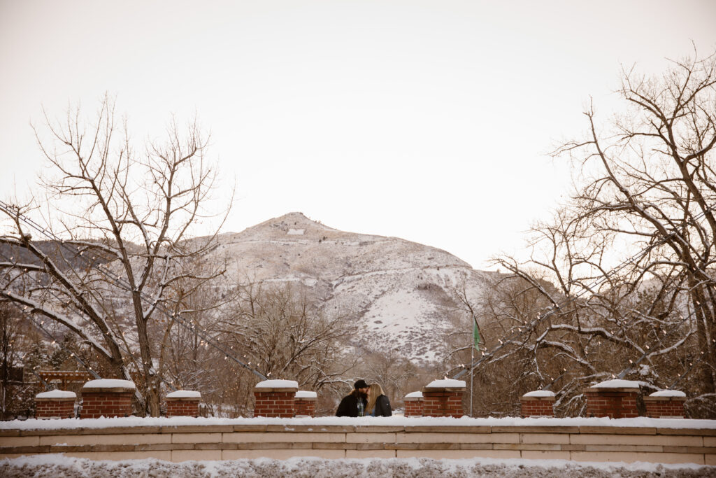 Couple cuddled together on a bridge over a river with a snowy mountain in the background