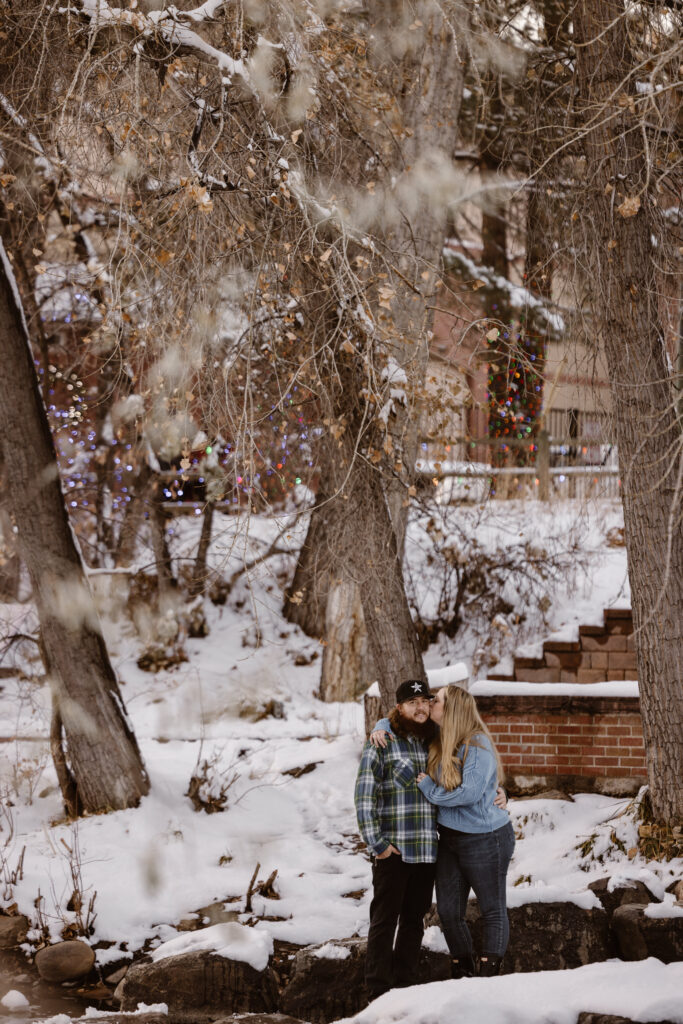 Couple cuddled together, woman giving her boyfriend a kiss on the cheek, next to a river with snow