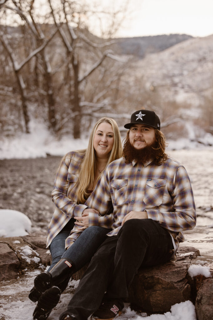 Couple sitting cuddled up next to each other next to a river with snow