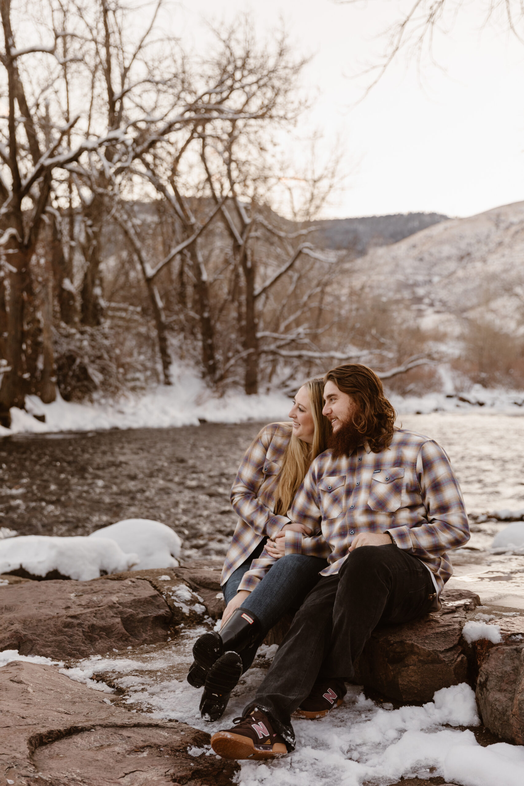 Couple sitting cuddled up next to each other next to a river with snow