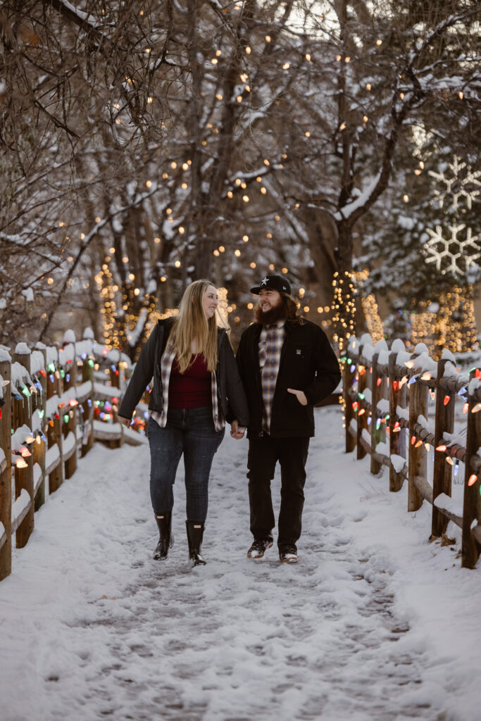 Couple walking hand in hand down a snowy sidewalk with Christmas lights all over