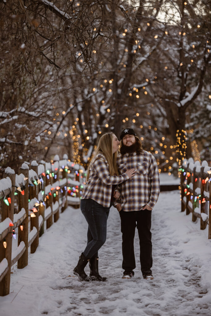 Couple on a snowy sidewalk with Christmas lights all over, woman looking at her boyfriend with her hand on his chest