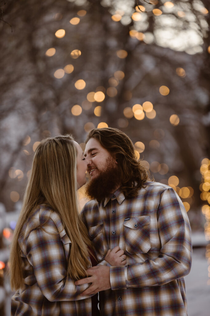 Couple going in for a kiss on a snowy sidewalk with Christmas lights in the background