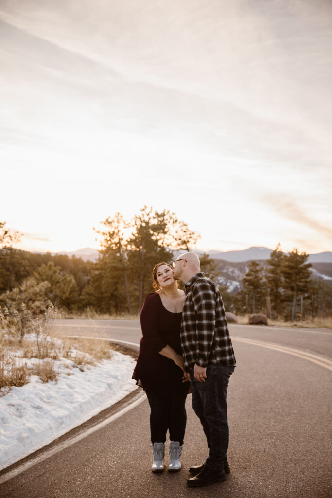 Man kissing his fiancé on the cheek on a mountain road at sunset