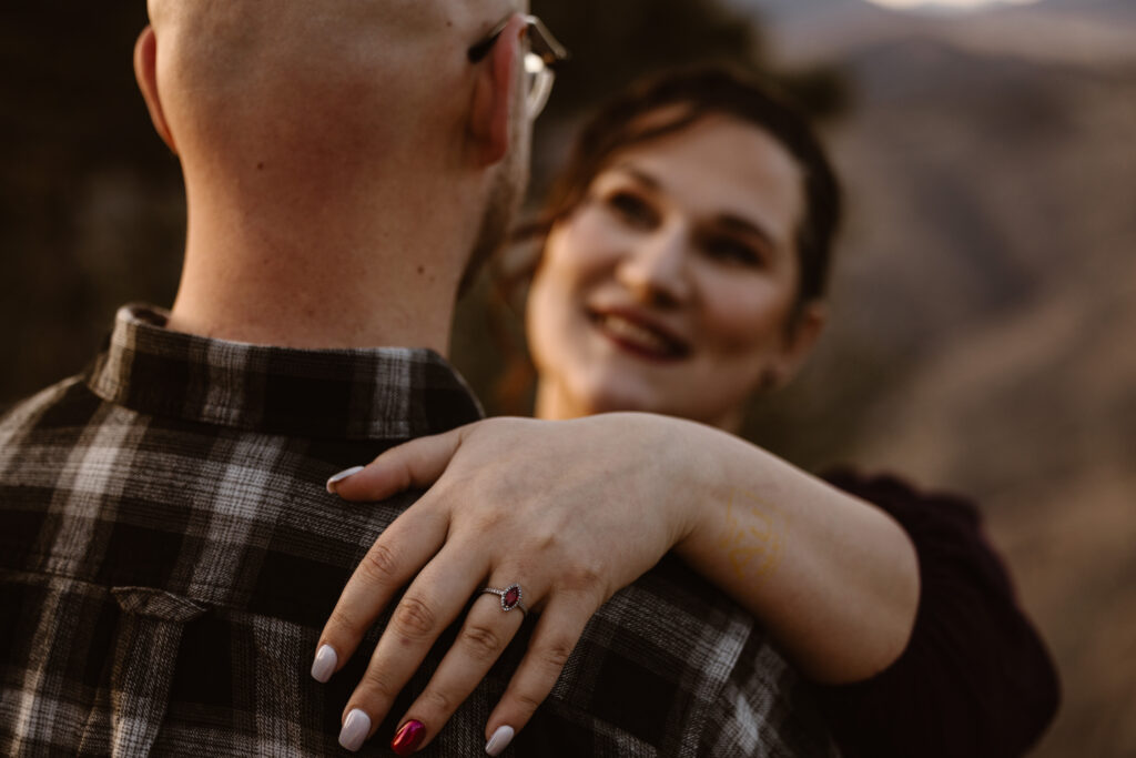 Woman's hand with her engagement ring on, with her looking at her fiancé blurred out in the background