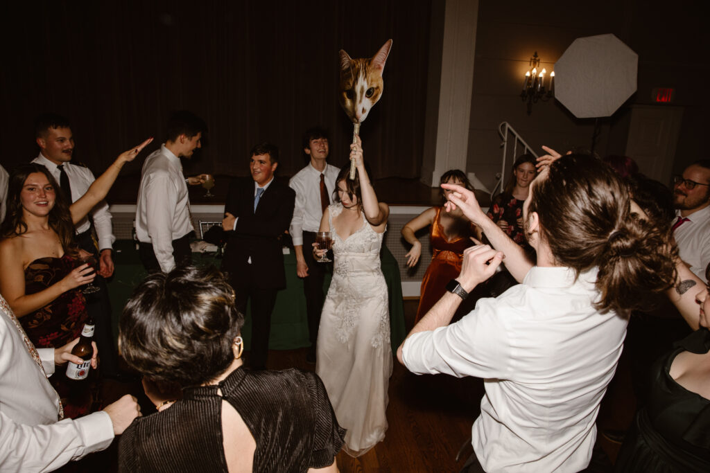 Bride holding oversized cat head cut out on a stick in a crowd of friends and family dancing