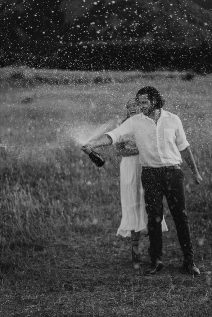 Black and white photo of a couple spraying champagne in an open field during their Boulder, Colorado engagement session.