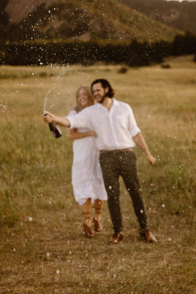 Blurred photo of a couple laughing and spraying champagne in a golden field during their Boulder engagement session, with mountain views in the background.