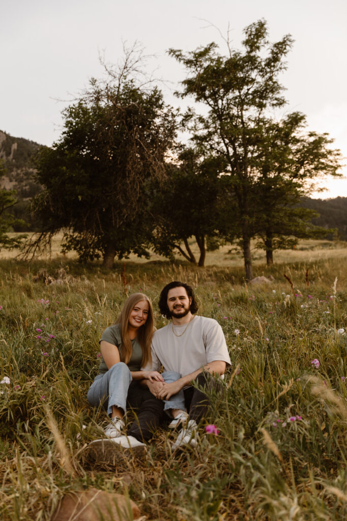 Couple sitting in a field of wildflowers at Chautauqua Park in Boulder, Colorado during their engagement session, smiling at the camera in casual outfits.