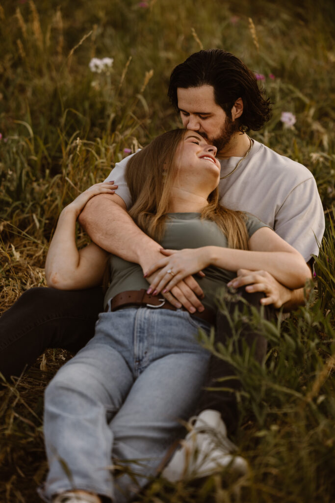 Engaged couple laying in a field at Chautauqua Park in Boulder, Colorado, with the groom-to-be kissing his fiancée’s forehead as she laughs.