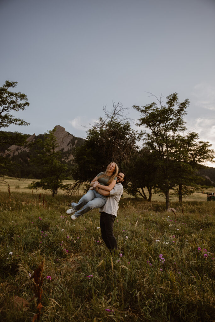 Playful engagement photo at Chautauqua Park in Boulder, Colorado, with the groom-to-be lifting his laughing fiancée in a wildflower field at sunset.