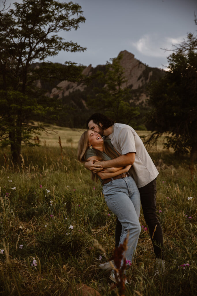 Playful engagement photo of a couple laughing and hugging in a wildflower meadow at Chautauqua Park, Boulder, Colorado with Flatirons in the background.