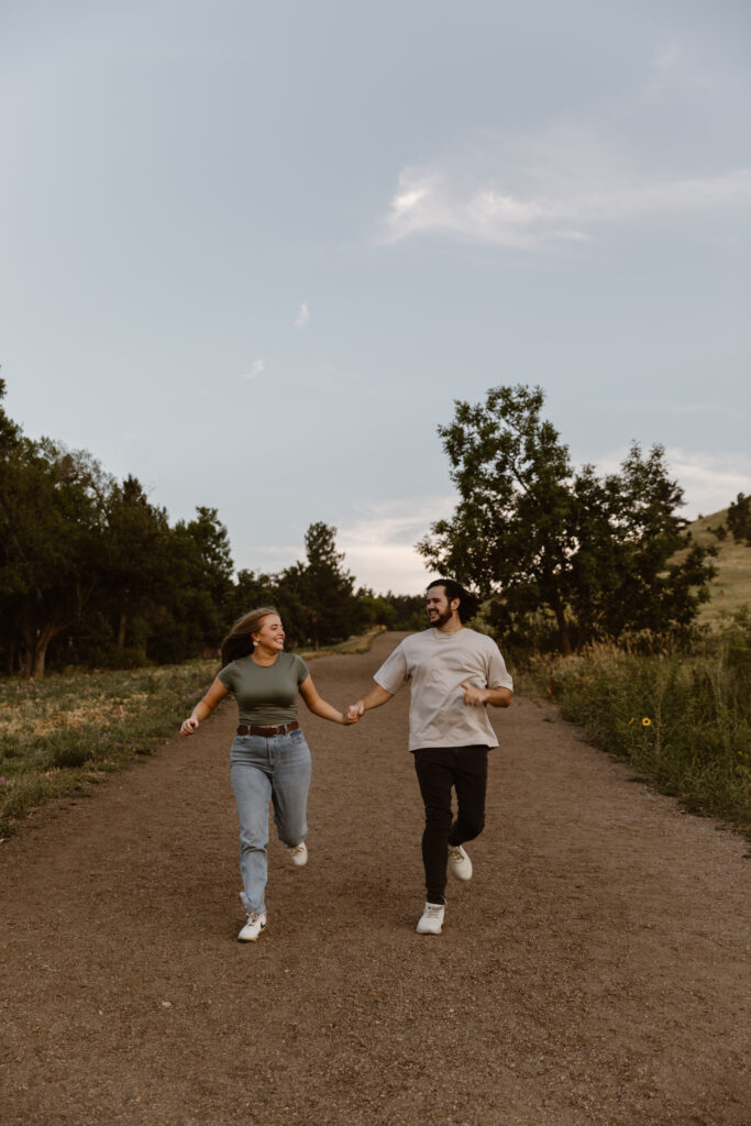 Engaged couple running hand in hand down a dirt path at Chautauqua Park in Boulder, Colorado, smiling joyfully during their engagement session.