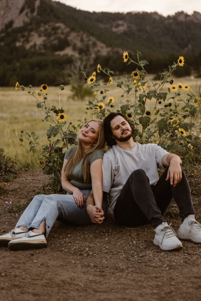 Couple sitting on a dirt path in front of wild sunflowers at Chautauqua Park in Boulder, Colorado, holding hands and leaning against each other during their engagement session.