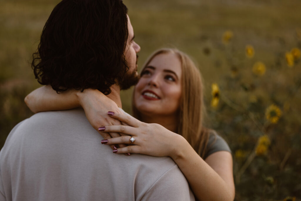 Close-up of a woman’s hand with an engagement ring resting on her partner’s shoulder as she smiles up at him during their Boulder engagement session.
