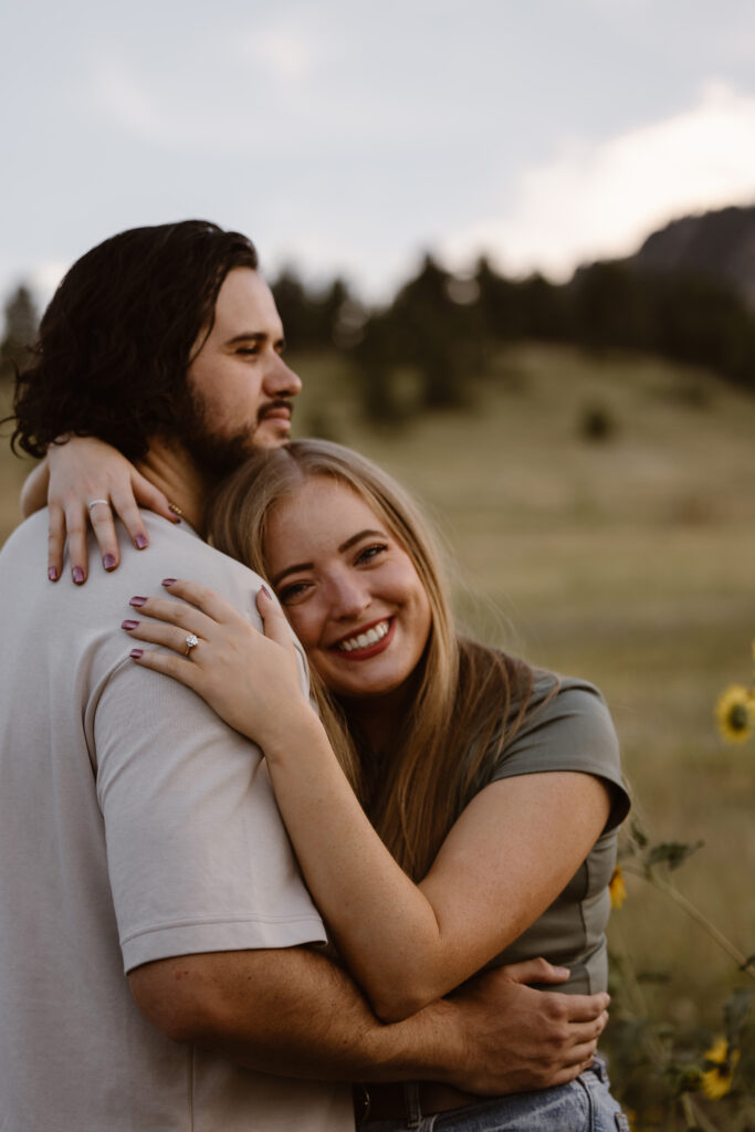 Woman smiling brightly at the camera while hugging her partner from behind, showing off her engagement ring during a Boulder engagement session.