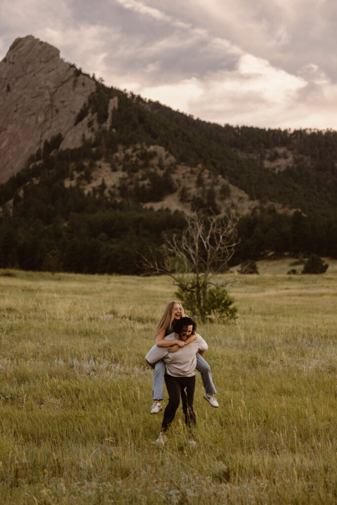 Woman laughing as her fiancé gives her a piggyback ride in an open meadow with the Flatirons in the background during a Colorado engagement session.