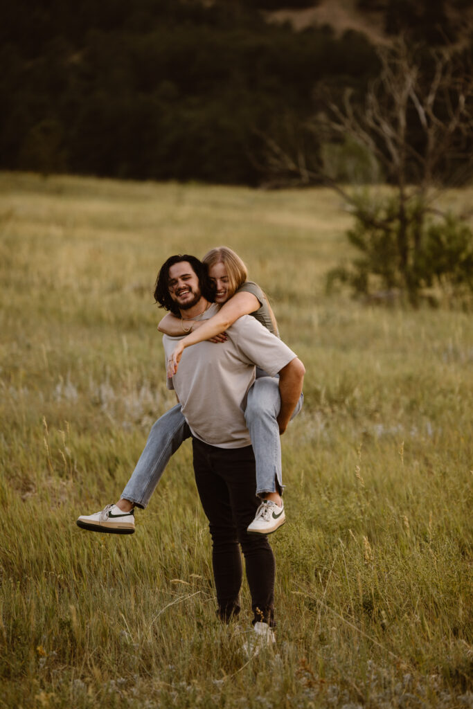 Couple laughing as the man gives his fiancée a piggyback ride through a grassy meadow during their Boulder, Colorado engagement session.