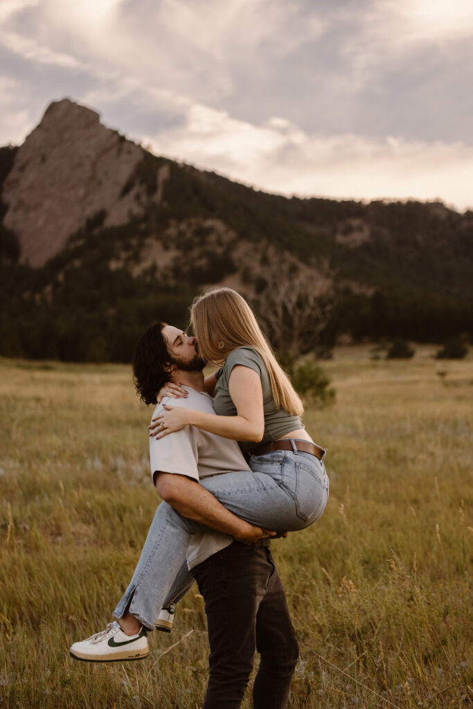 Playful engagement photo of a couple kissing while the man lifts his fiancée in a meadow near the Flatirons in Boulder, Colorado.