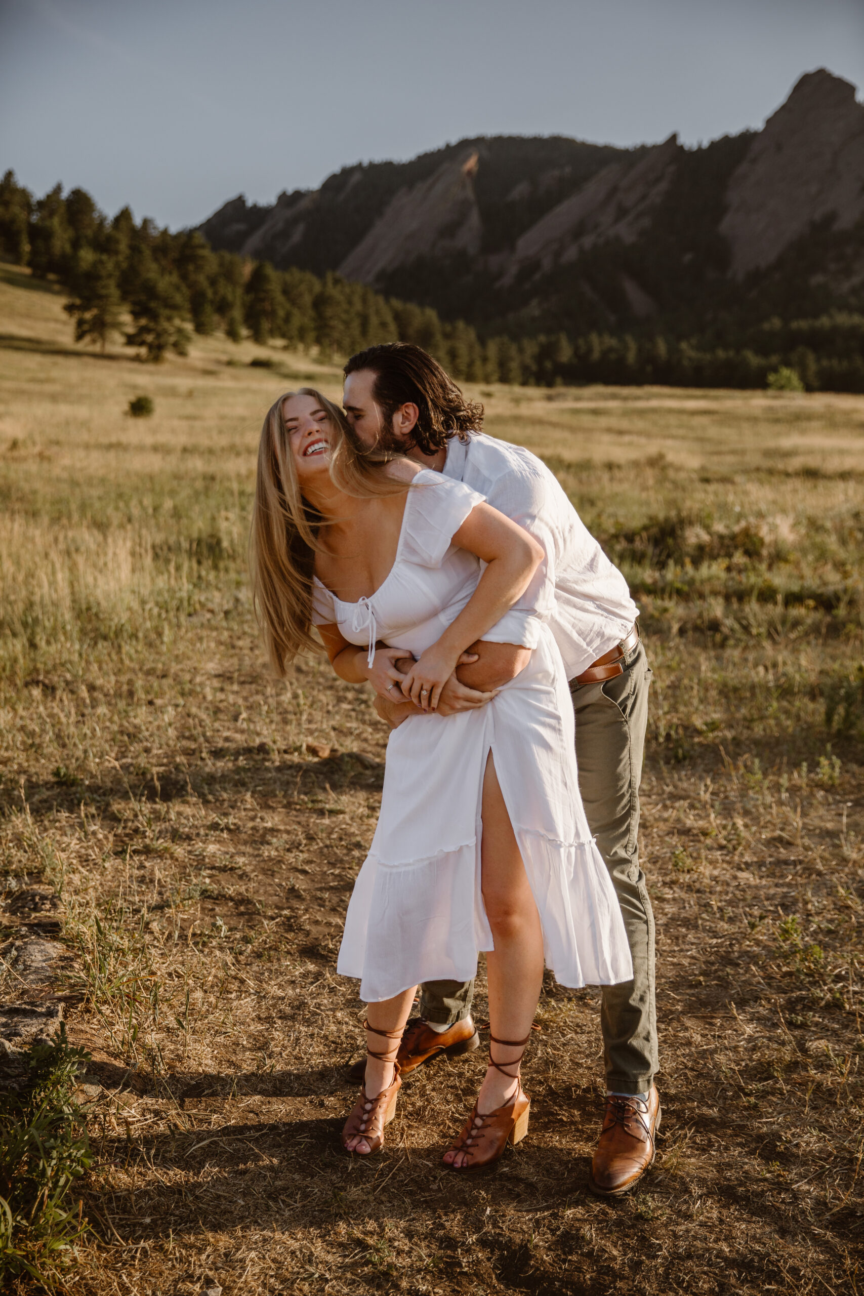 Couple laughing and embracing during a playful engagement photoshoot at Chautauqua Park in Boulder, Colorado with the Flatirons in the background.
