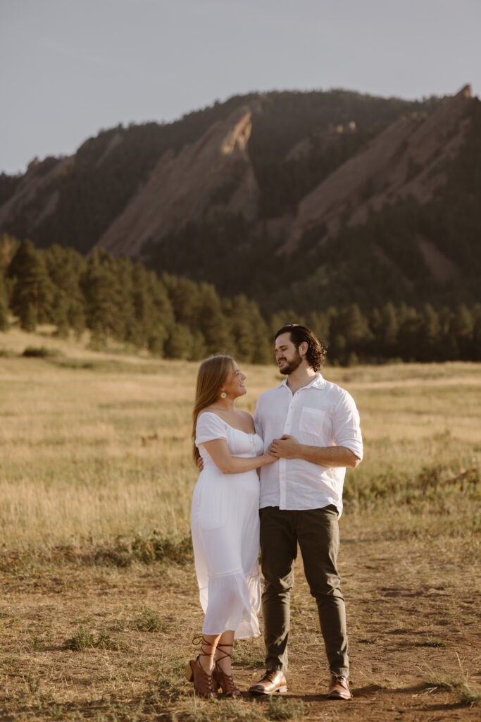 Couple standing together in a sunlit field at Chautauqua Park in Boulder, Colorado, with the Flatirons in the background during their engagement photo session.