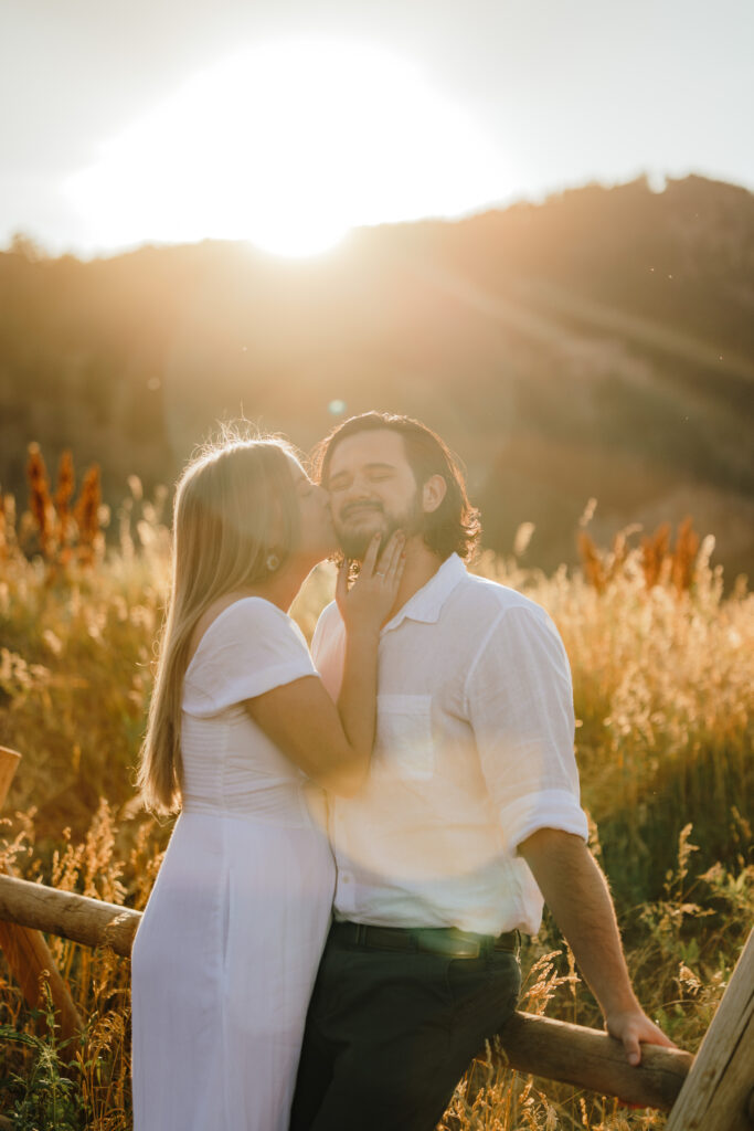 Woman kissing her fiancé’s cheek during a romantic engagement session in Boulder, Colorado, with golden sunlight streaming over the Flatirons.