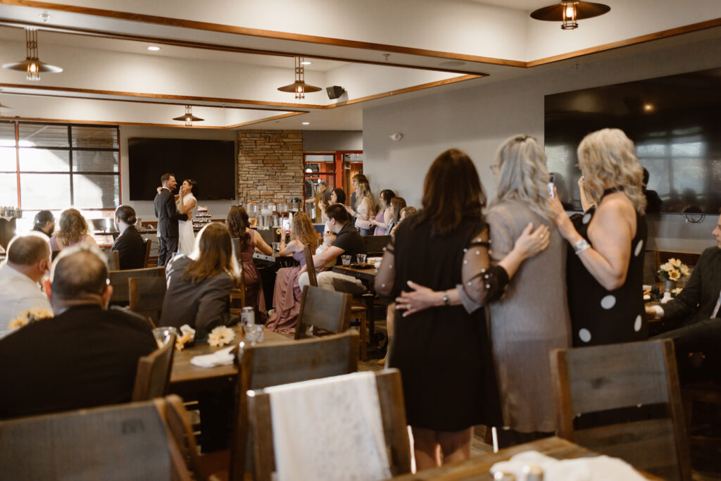 Caitlin’s mom and friends embrace while watching bride and groom’s first dance at Walnut Creek Golf Preserve wedding reception in Westminster Colorado by Colorado wedding photographer.