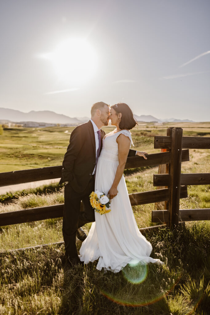 Bride and groom sharing a romantic kiss at sunset by a wooden fence at Walnut Creek Golf Preserve wedding in Westminster Colorado with mountain views by Colorado wedding photographer.