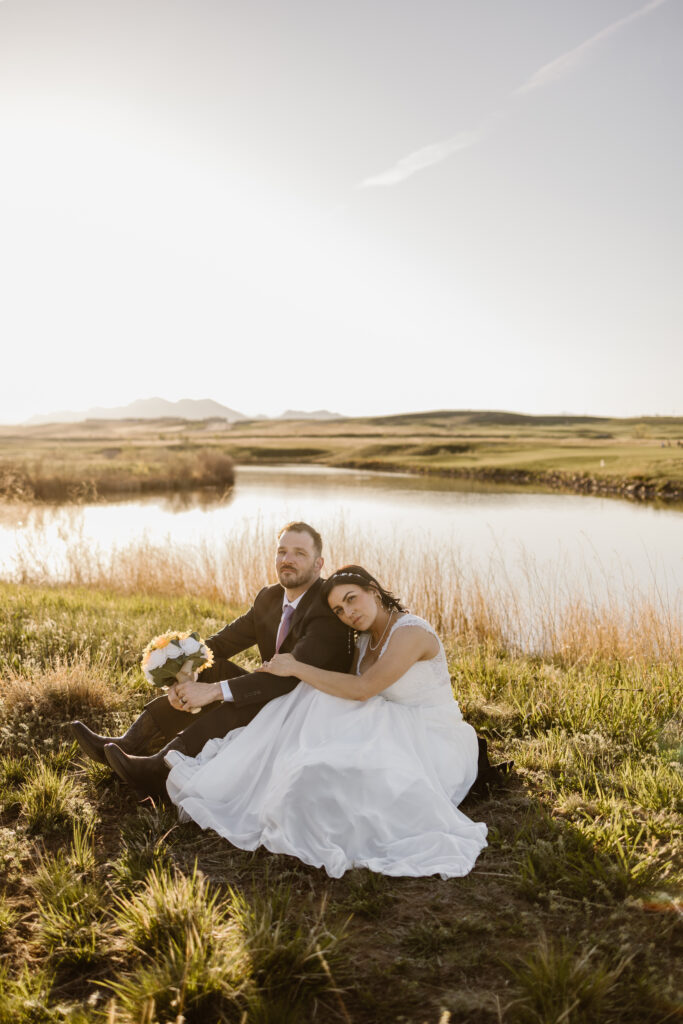 Bride and groom sitting together by a pond during golden hour portraits at Walnut Creek Golf Preserve wedding in Westminster Colorado captured by Colorado wedding photographer.
