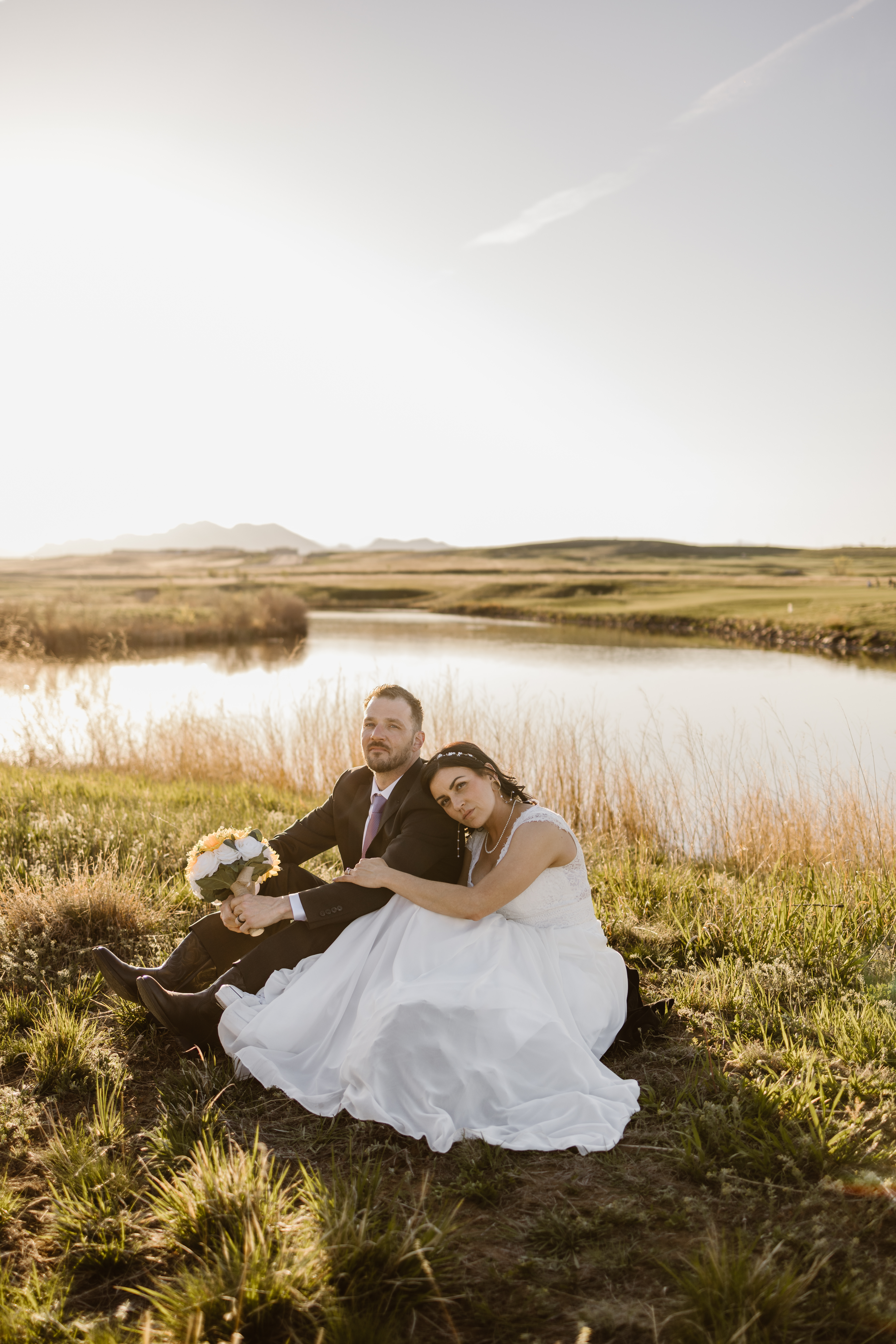 Bride and groom sitting together by a pond during golden hour portraits at Walnut Creek Golf Preserve wedding in Westminster Colorado captured by Colorado wedding photographer.