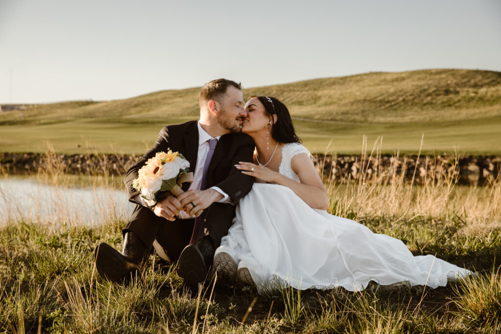 Bride and groom sharing a kiss while sitting in the grass during golden hour portraits at Walnut Creek Golf Preserve wedding in Westminster Colorado captured by Colorado wedding photographer.